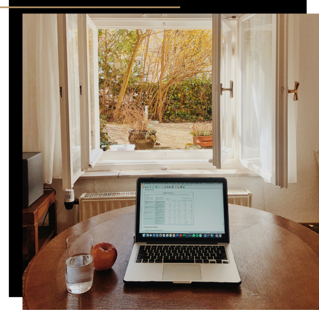 A laptop on a table next to an apple and a glass of water in front of an open window