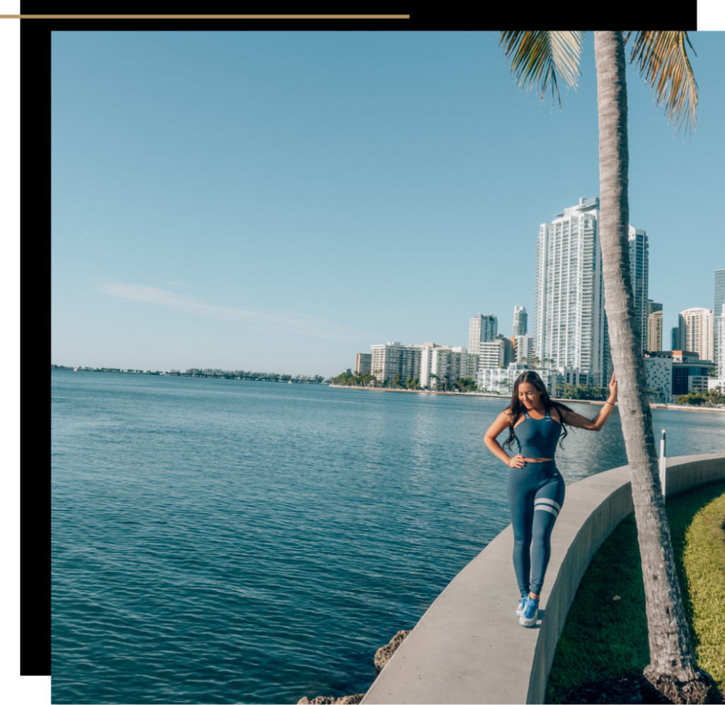 Isabella in activewear touching a palm tree by the water in Miami